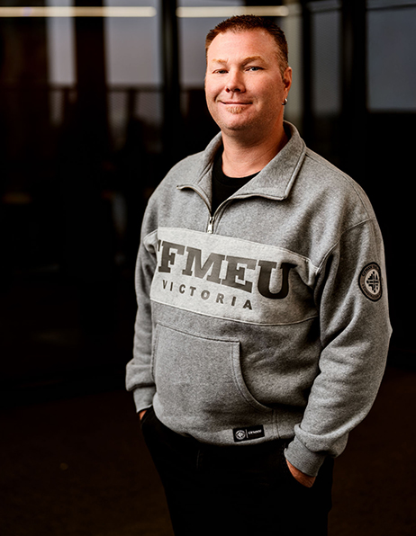 Man standing indoors wearing a gray FMEU Victoria hoodie with hands in pockets.