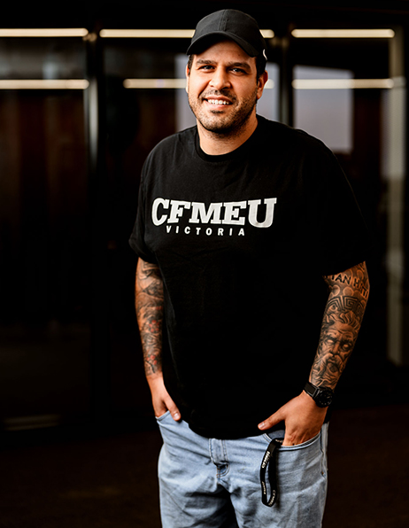 Man wearing a black CFMEU Victoria T-shirt and cap, standing with hands in pockets, visible tattoos on both forearms.