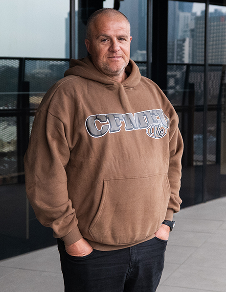 Man wearing a brown hoodie with a large logo on the chest, standing with hands in pockets outside a glass building.