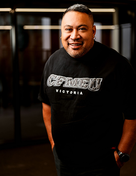 Smiling man wearing a black CEMEU Victoria t-shirt, standing indoors near glass doors.