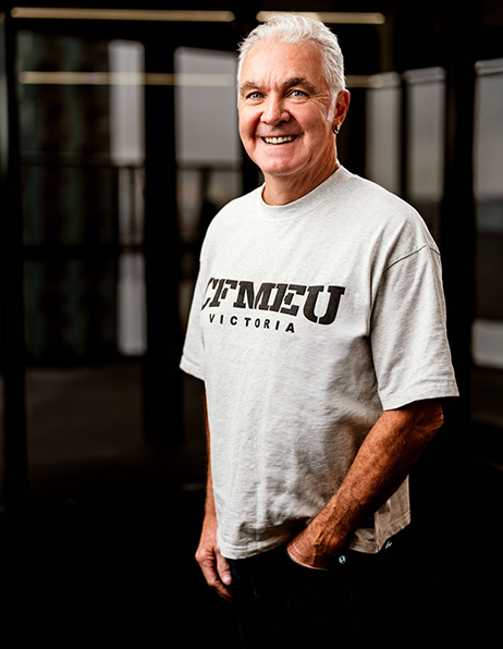 Smiling older man in a light gray T-shirt that reads TFMEU Victoria, standing in a gym area.
