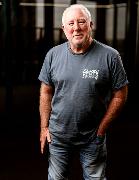 Older man with white hair and beard wearing a gray t‑shirt, standing in a dark indoor space with his hands in his pockets.