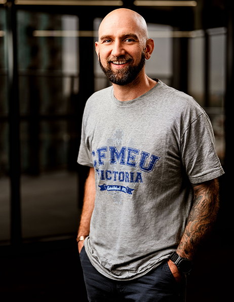 Portrait of a smiling, tattooed bald man with a beard, wearing a gray T-shirt with a blue graphic, standing indoors with hands in pockets.