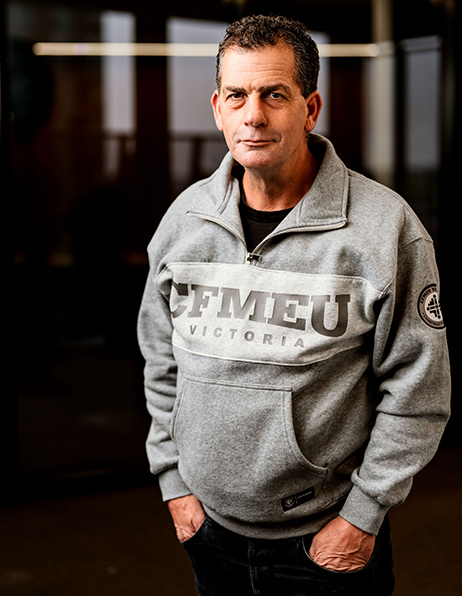 Portrait of a man standing indoors with hands in pockets, wearing a gray CFMEU Victoria sweatshirt.