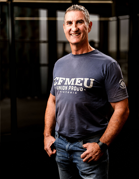 Portrait of a smiling, middle-aged man wearing a dark blue CFMEU 'Union Proud' T-shirt and jeans, standing with hands in pockets.