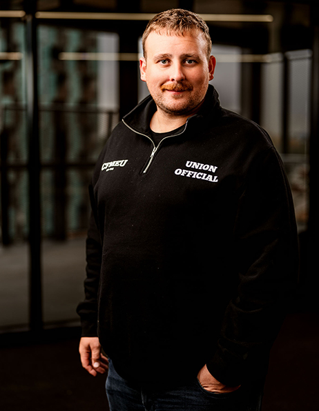 Portrait of a man wearing a black zip-neck sweatshirt with 'Union Official' on the chest, standing indoors.