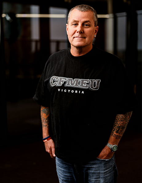 Man with short gray hair and tattooed arms wearing a black CFMEU Victoria t-shirt, standing with hands in pockets in a dim indoor setting.