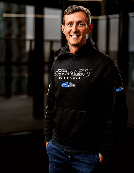 Smiling man wearing a black CFMEU Victoria hoodie, standing with hands in pockets in a dim indoor space.