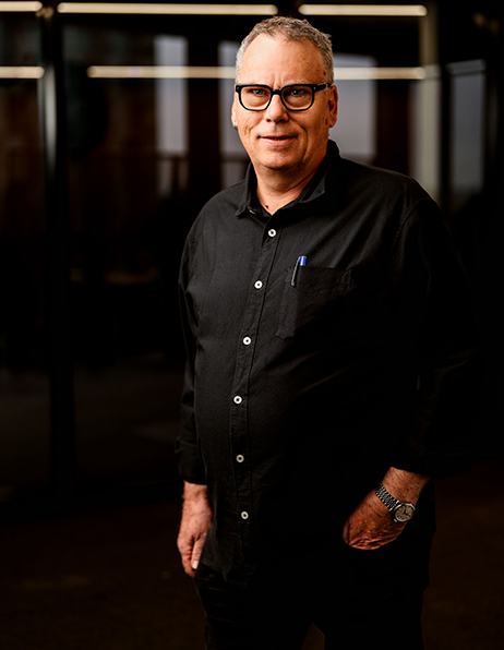 Portrait of a middle-aged man with gray hair and glasses, wearing a black button-down shirt, standing in a dim office setting with hands in pockets.