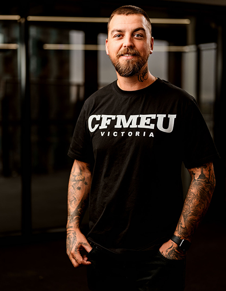 Man with a beard and tattoos wearing a black CFMEU Victoria t-shirt, standing in a gym with hands in pockets and a smile.