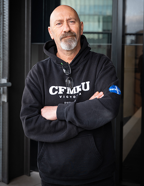 Man with a gray goatee and crossed arms wearing a black CFMEU hoodie, standing at a building entrance.