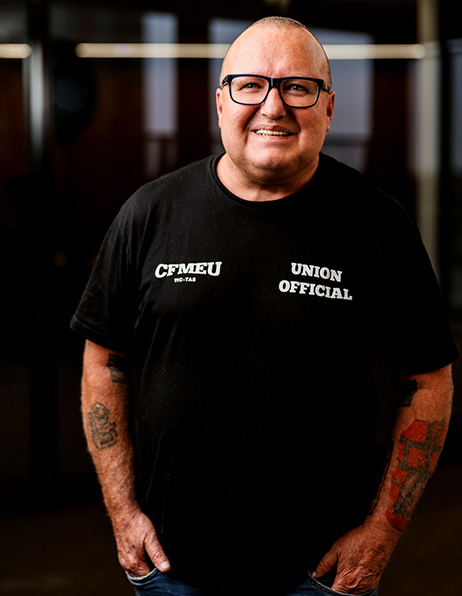 Smiling bald man with glasses in a black T-shirt that reads 'UNION OFFICIAL' and 'CFMEU,' standing indoors with tattooed arms.