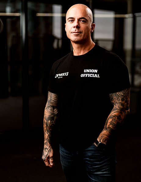 Tattooed, bald man in a black T‑shirt reading 'UNION OFFICIAL' stands in a dim gym‑like space.