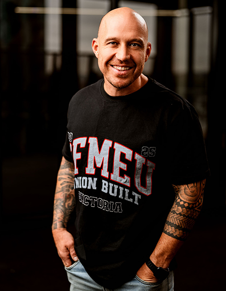 Smiling bald man in a black graphic T-shirt with bold white and red text, standing with hands in pockets in a dark indoor setting.
