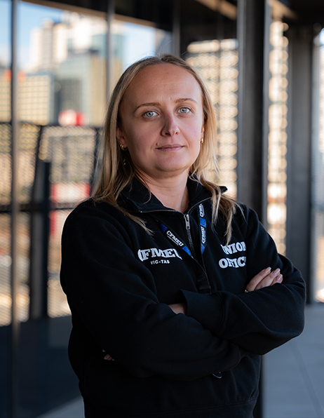 Woman with blonde hair standing outdoors with arms crossed, wearing a black hoodie and a lanyard with badges, city buildings in the background.