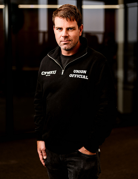 Man standing indoors, wearing a black zip-up sweater that reads 'CFMEU UNION OFFICIAL' on the chest.