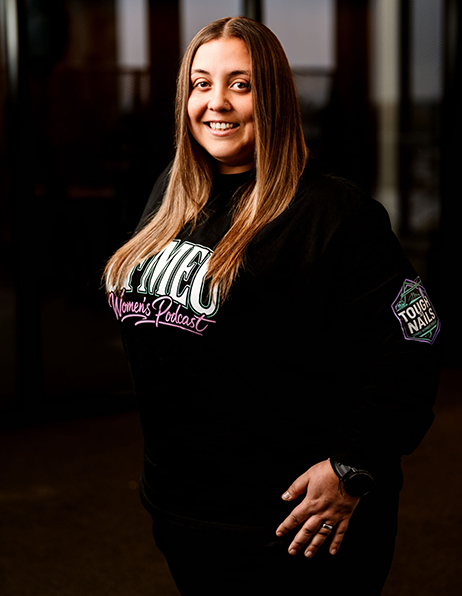 Smiling woman in a black sweatshirt with a 'WOMEN'S Podcast' logo, standing in a dim indoor setting; patch visible on the left sleeve.