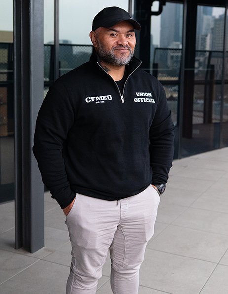 A union official posing outdoors, wearing a black zip‑neck pullover with CFMEU and 'Union Official' logos, hands in pockets.