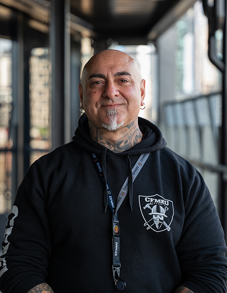 Smiling bald man with a goatee and earrings, wearing a black hoodie and a lanyard, standing in a sunlit indoor corridor.
